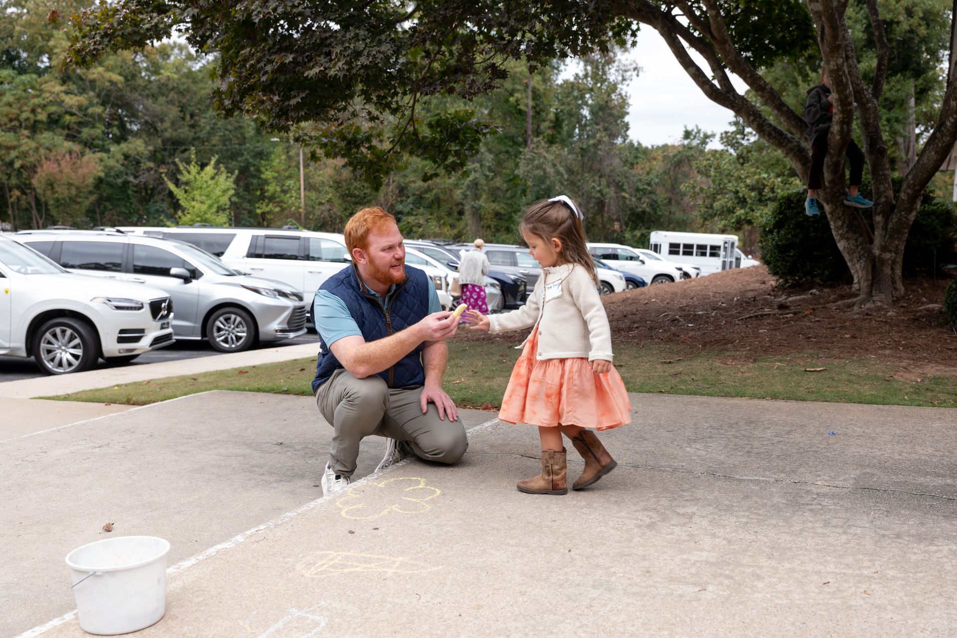Man kneels on a sidewalk handing an item to a girl in a peach dress near parked cars and trees.