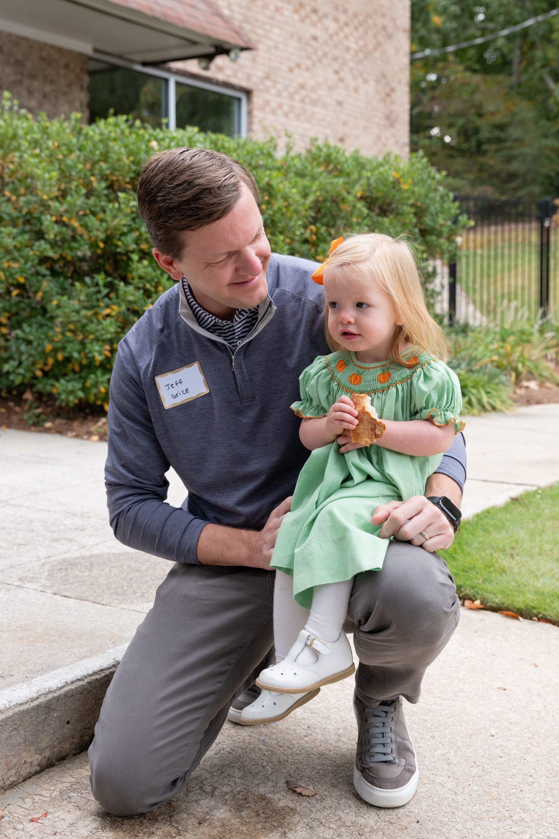 Man kneels beside a toddler on a sidewalk, smiling at her as she holds a small bouquet.