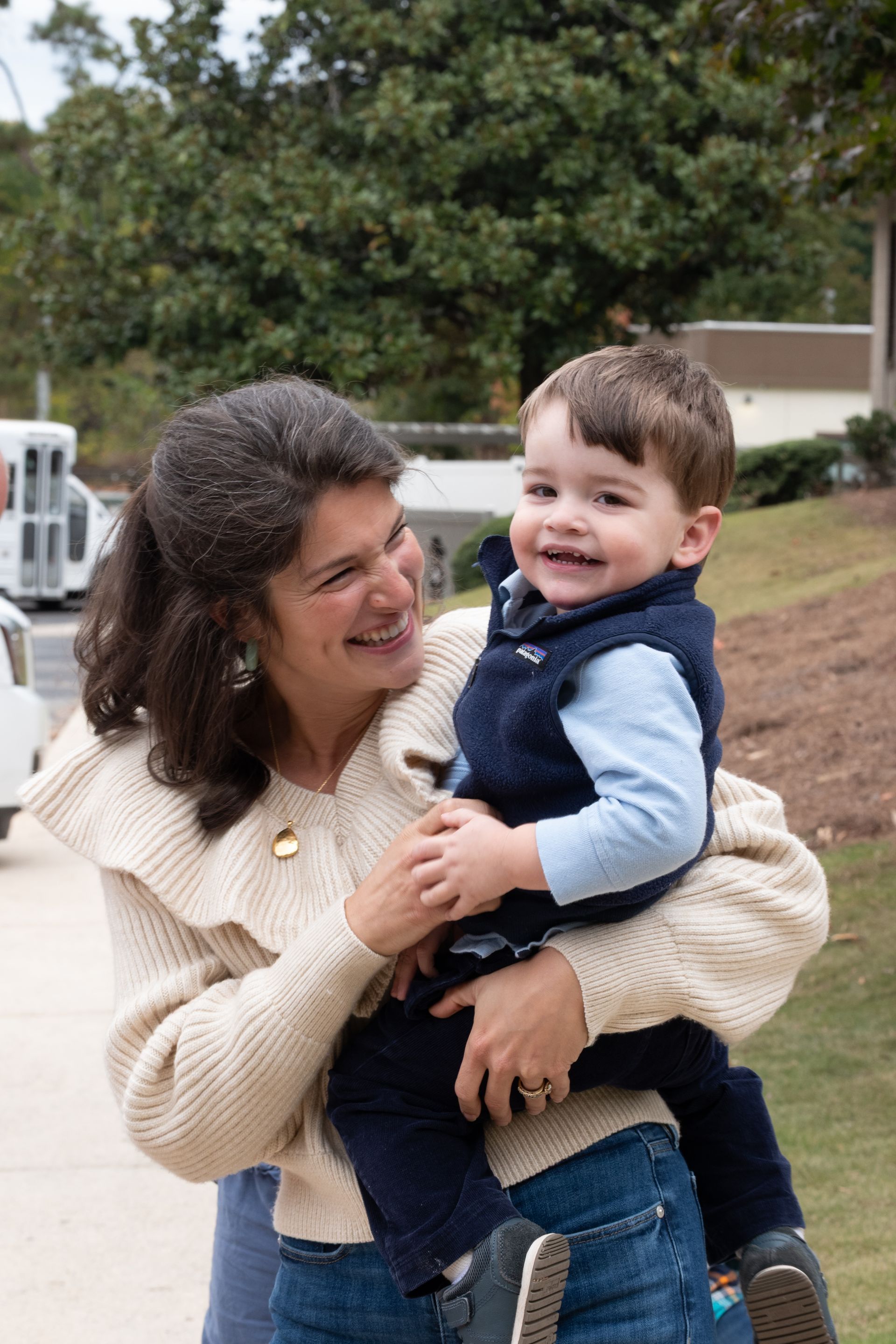 Woman holding smiling child outdoors near trees and parked cars