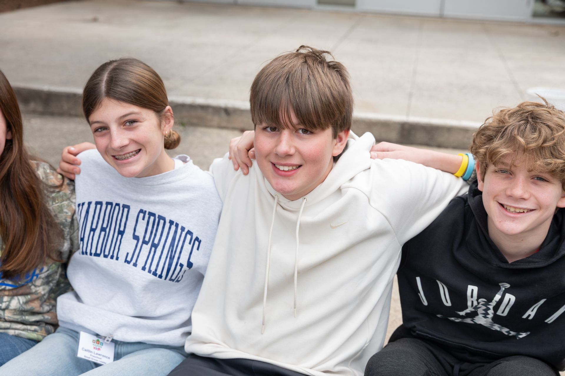 Three smiling friends sitting close together, wearing hoodies and sweaters outdoors on steps