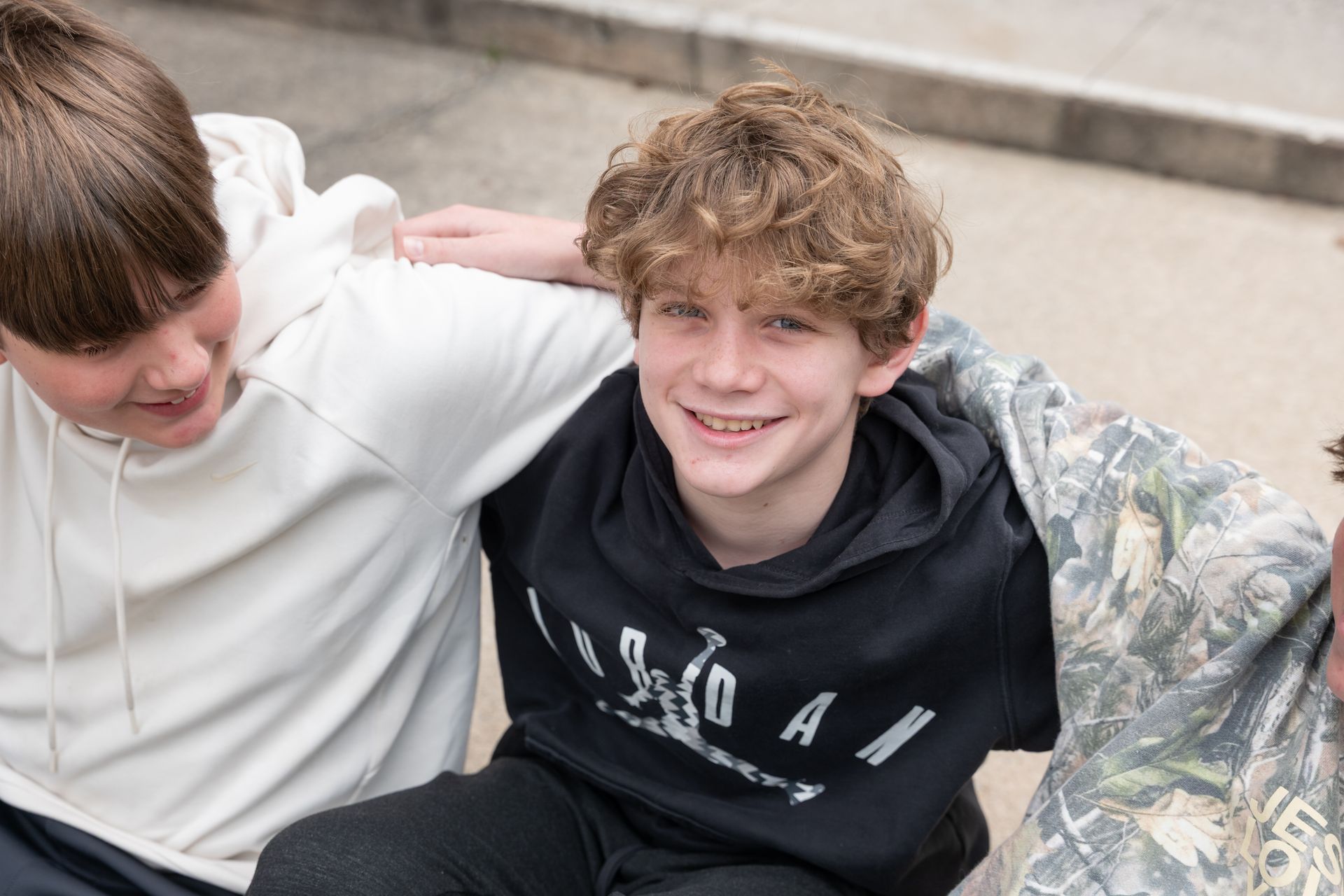 Two friends sitting close together outdoors, smiling at the camera; one in a white hoodie, one in a black hoodie.