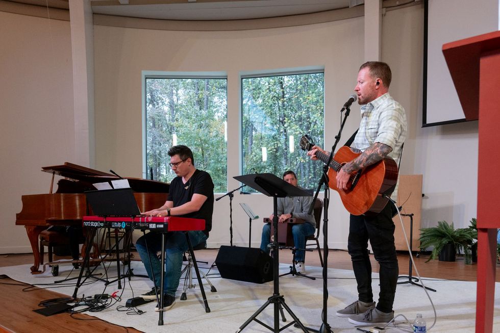 Musicians performing on a stage, with piano, keyboard, and guitar in a bright room with large windows
