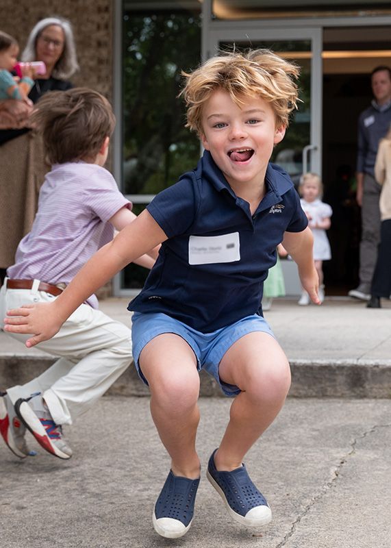 Smiling child in navy shirt and shorts crouches mid-jump on a sidewalk outside a building.