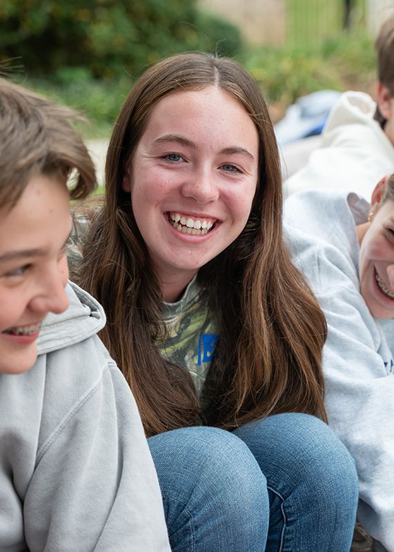 Smiling teen in a gray hoodie and jeans sitting outdoors with friends, one holding a mug.