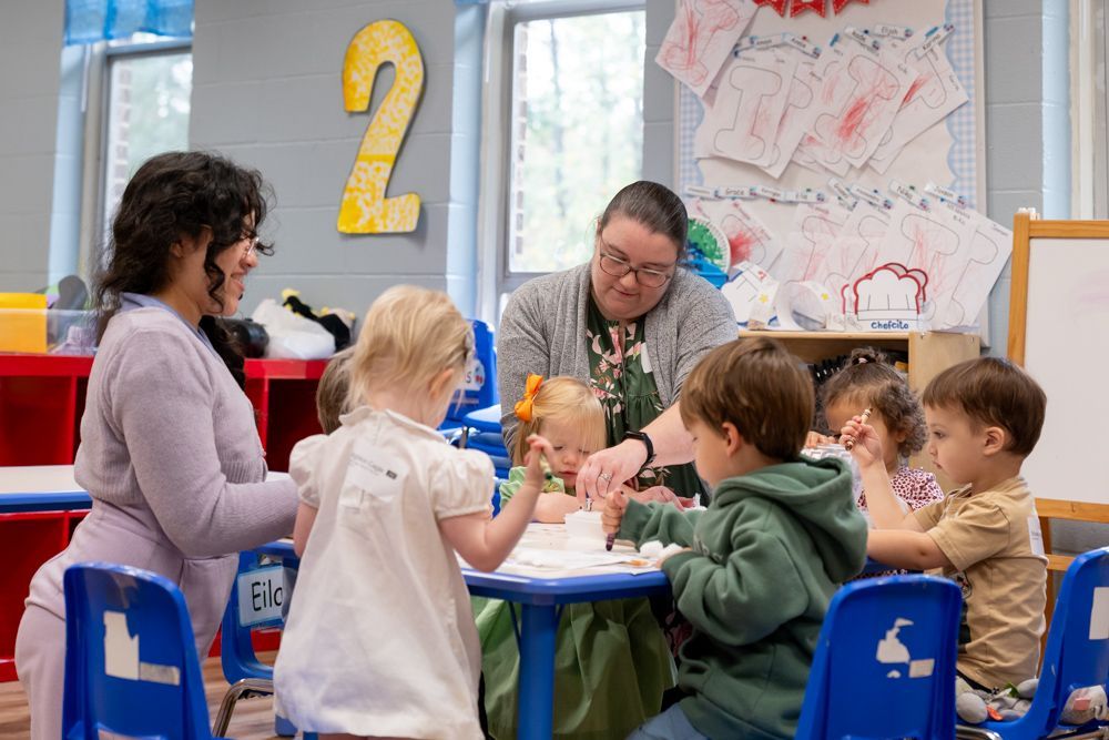 Teacher and children gathered around a table in a colorful classroom activity center.