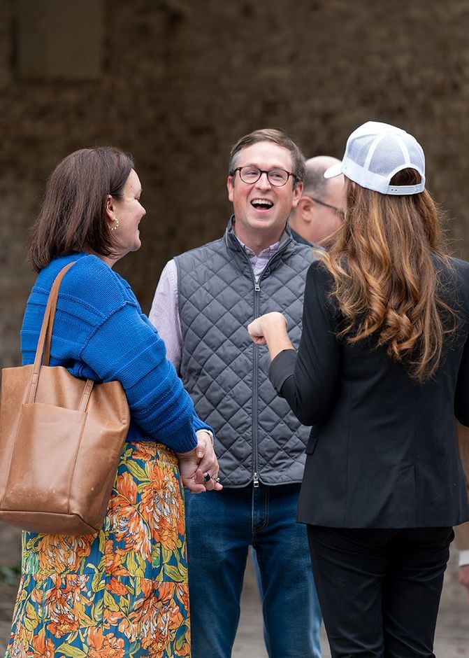 Three people chatting outdoors, with one man smiling in a gray quilted vest.
