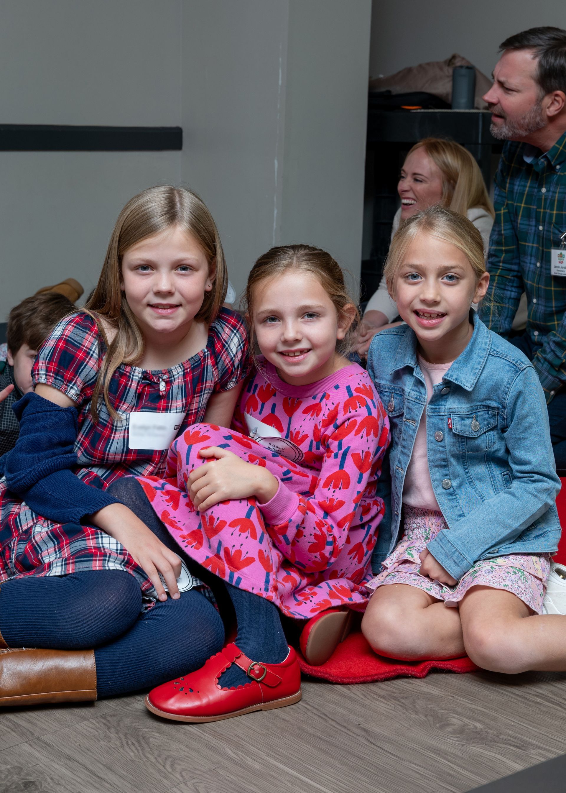 Three smiling children seated indoors, with adults standing behind them.