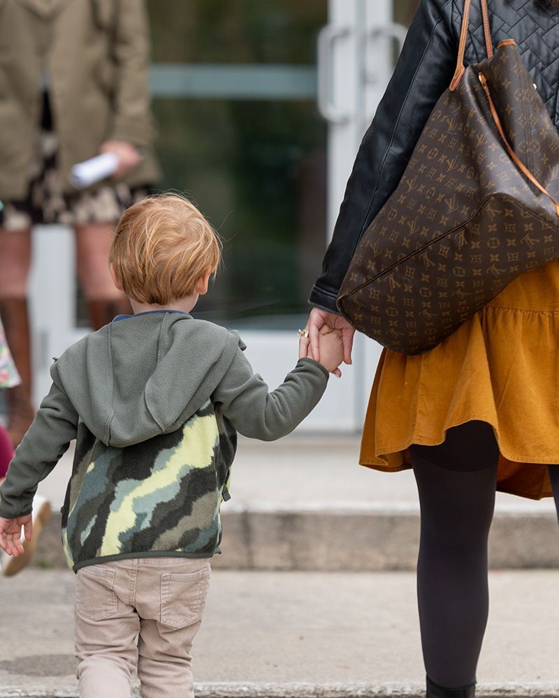 Adult and child walking hand in hand outdoors, seen from behind; child wears a green hoodie.