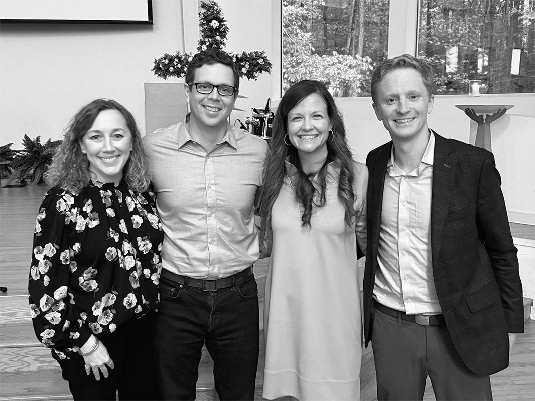 Four people standing together indoors, smiling at the camera in a casual group photo.