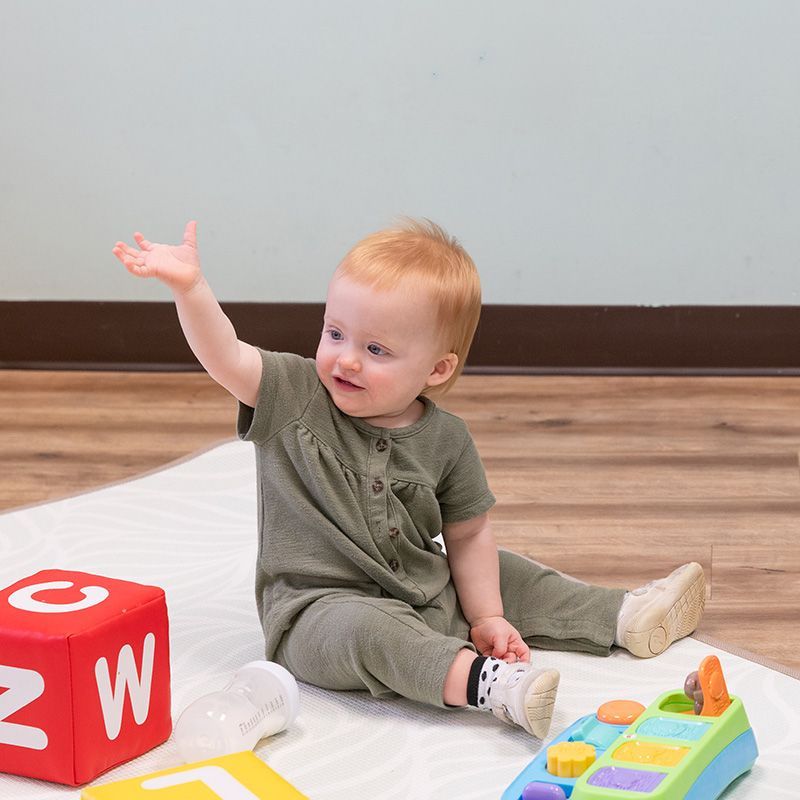 Toddler sitting on a white blanket, reaching up beside a red block and a colorful toy tray on a wooden floor