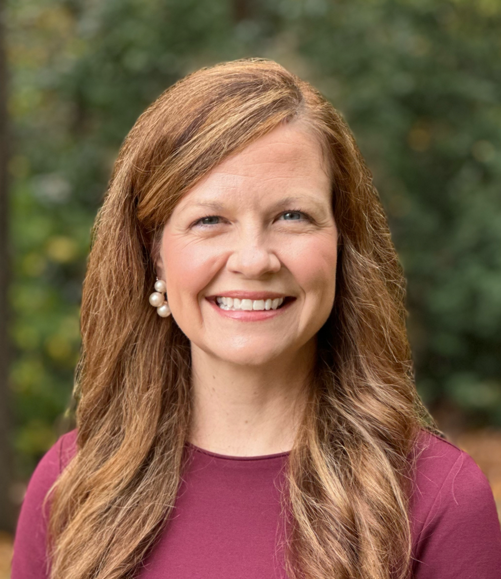 Lyndsay smiling with long wavy hair wearing a maroon top against a blurred outdoor background