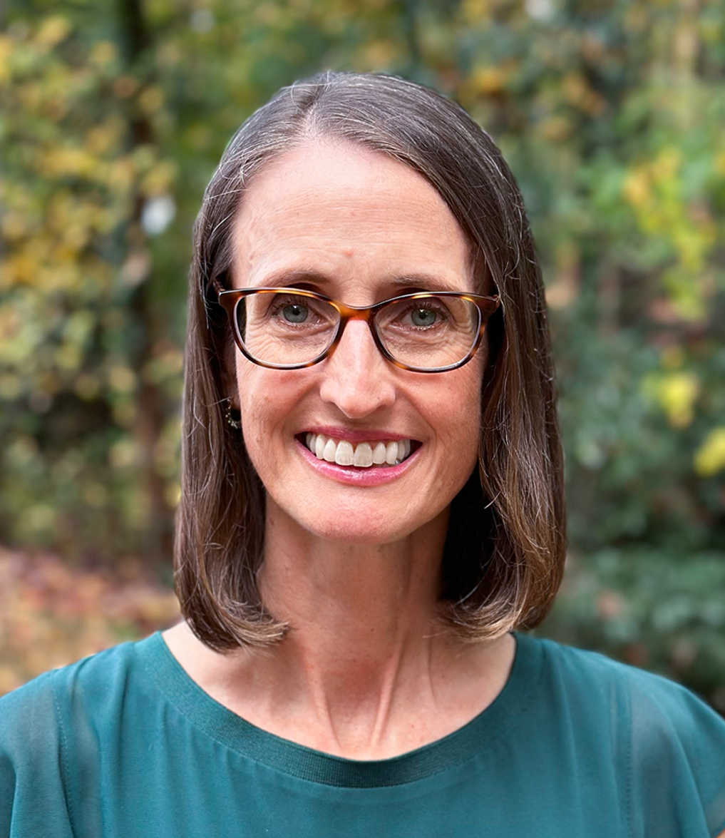 Miriam smiling with glasses and shoulder-length hair, wearing a teal top against a blurred outdoor background