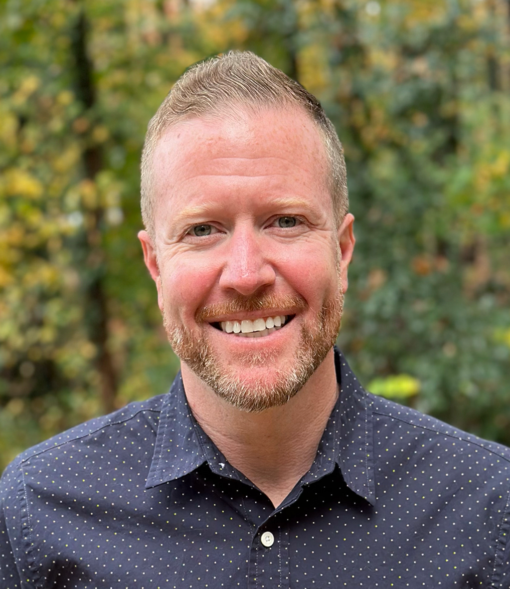 Ryan Horse smiling in a dark patterned shirt against a blurred outdoor background