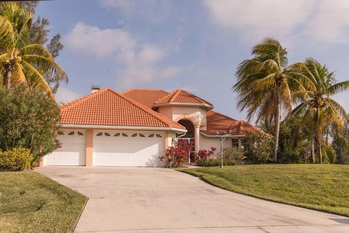 A beige house with an orange tile roof, white garage doors, palm trees, and a concrete driveway.