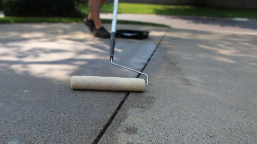 Person rolling sealant onto a concrete driveway crack with a roller and extended handle.