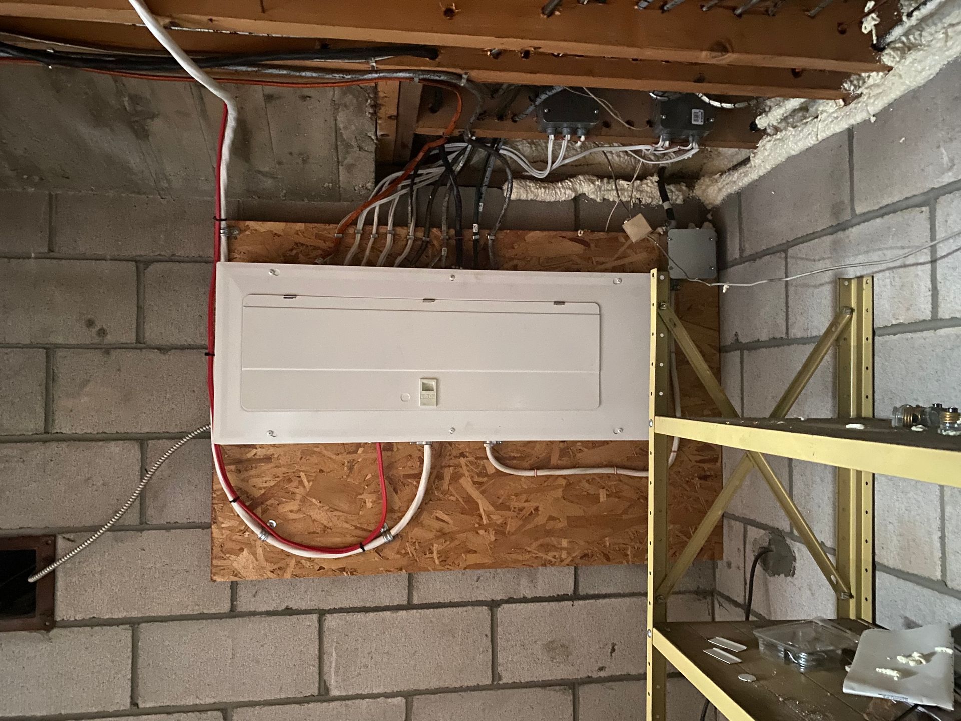 Electrical panel mounted on a wood panel in a cinder block basement. Wires and shelving present.