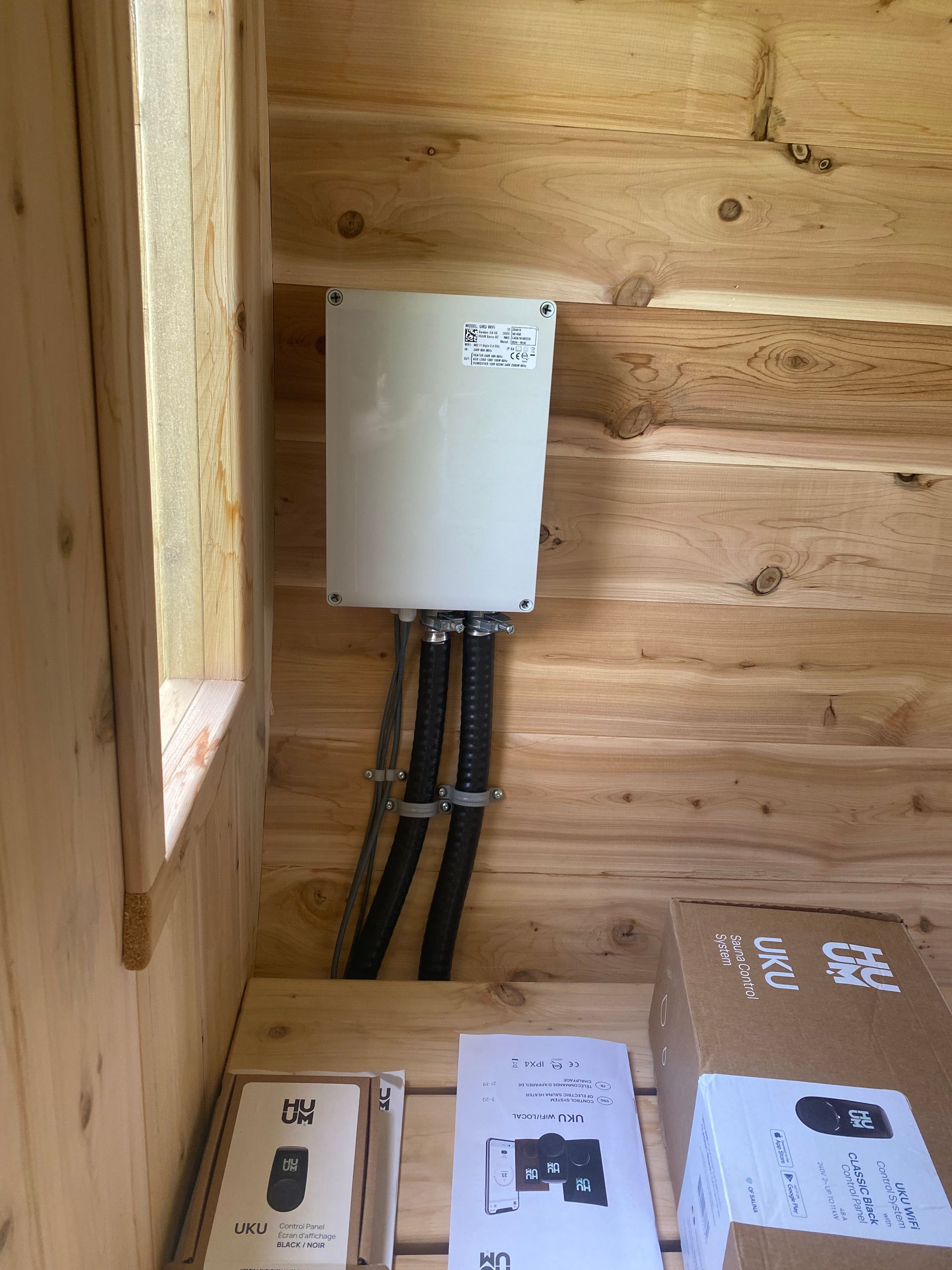 White electrical box mounted on a wooden wall with black conduits running down; paperwork and boxes sit below.