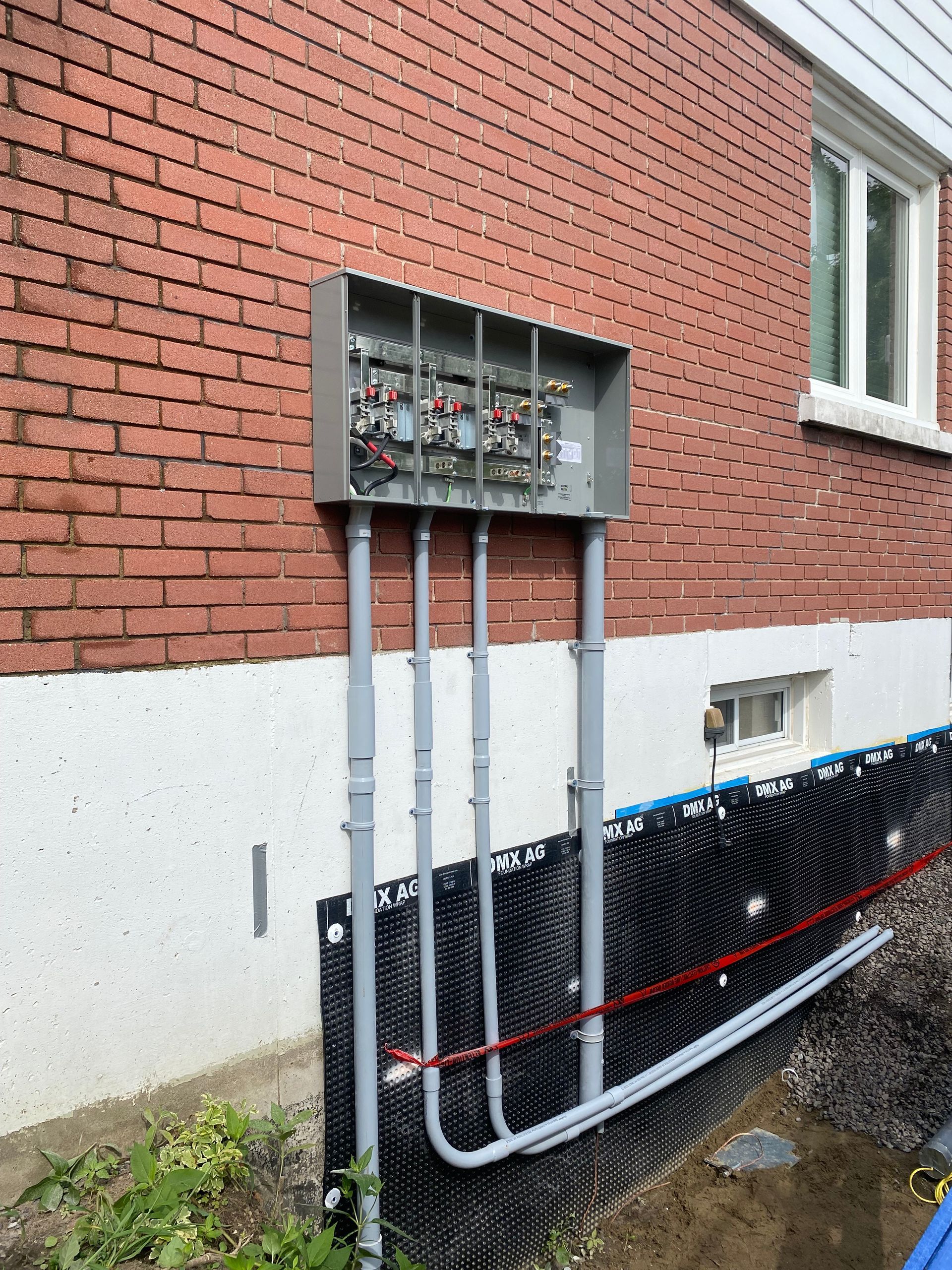 Electrical panel mounted on brick wall with conduit pipes, near a window and foundation protection.