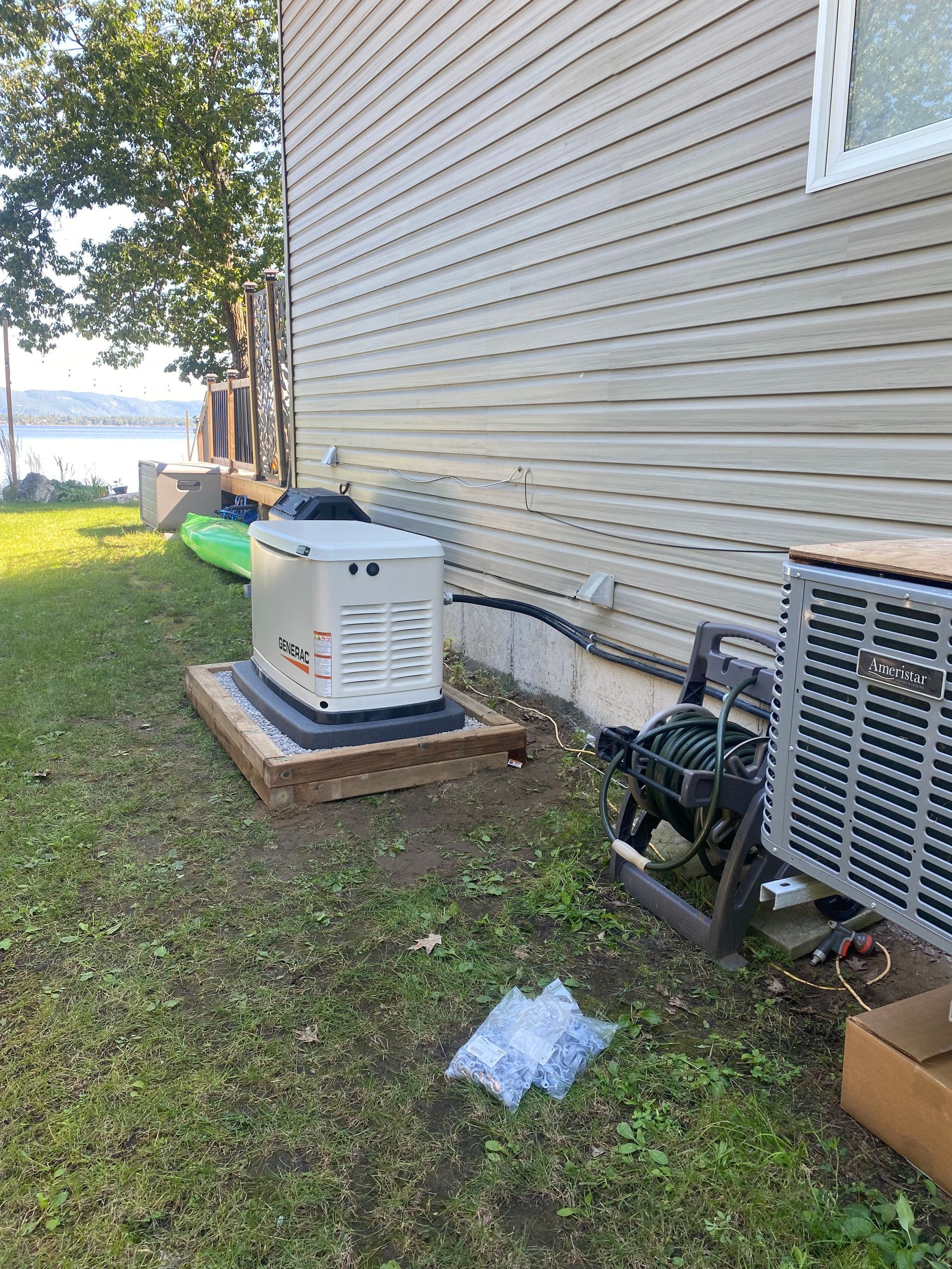 Generator on wooden platform next to house with AC unit, water hose, and view of water.