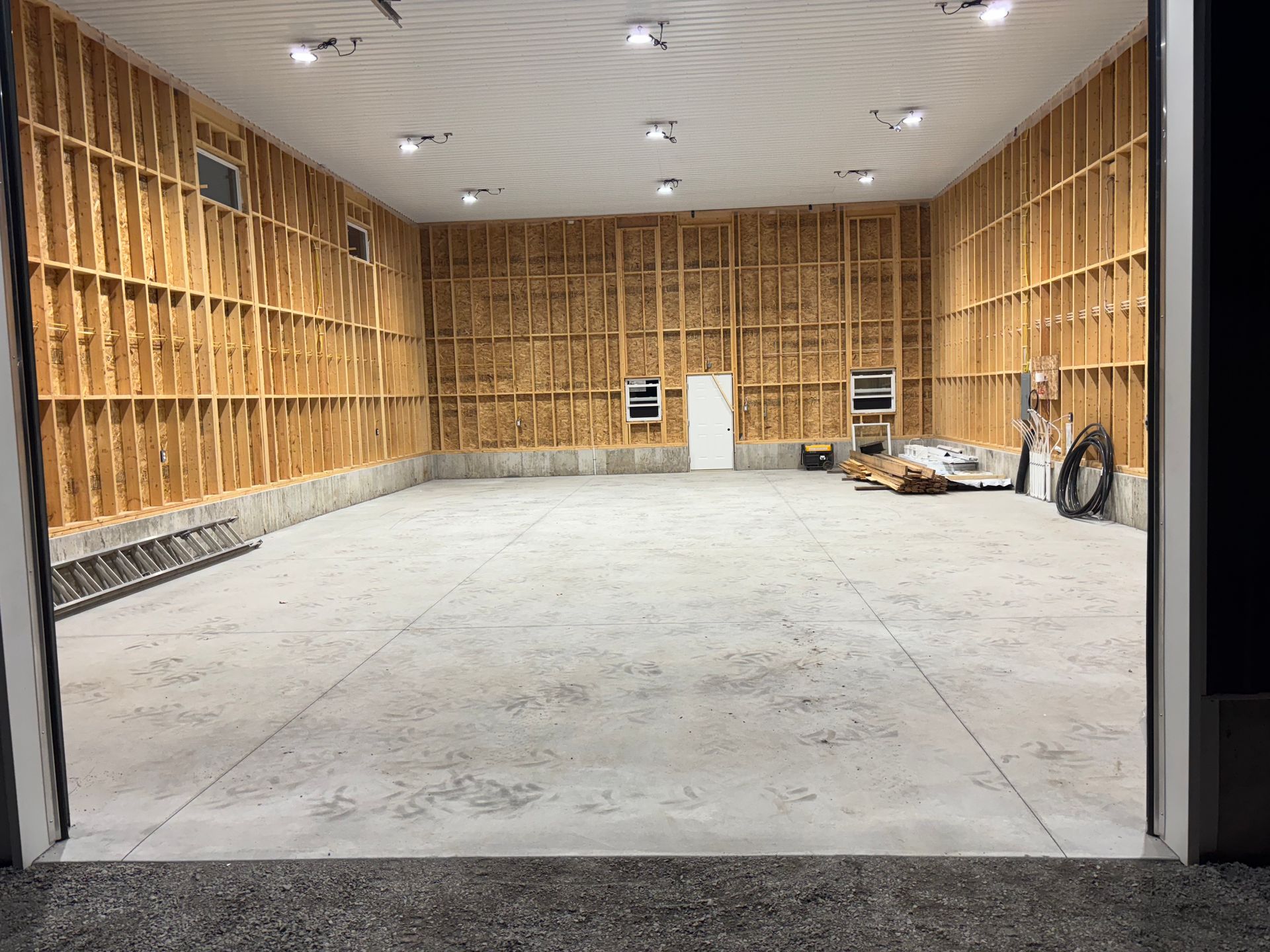 Interior view of a new, empty garage with concrete floor, exposed wooden framing, and bright overhead lights.