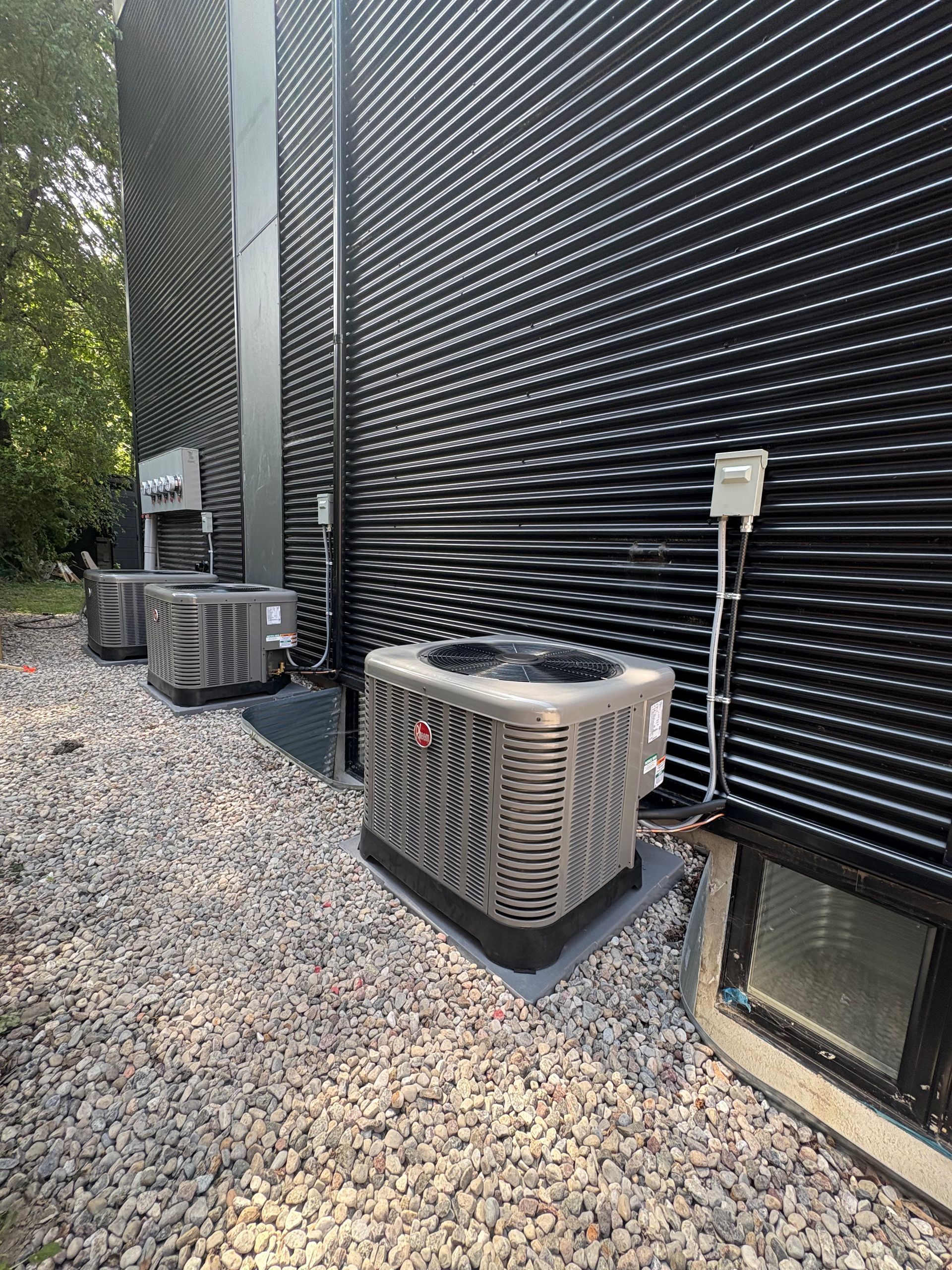 Three air conditioning units outside a building with corrugated black siding, on gravel.