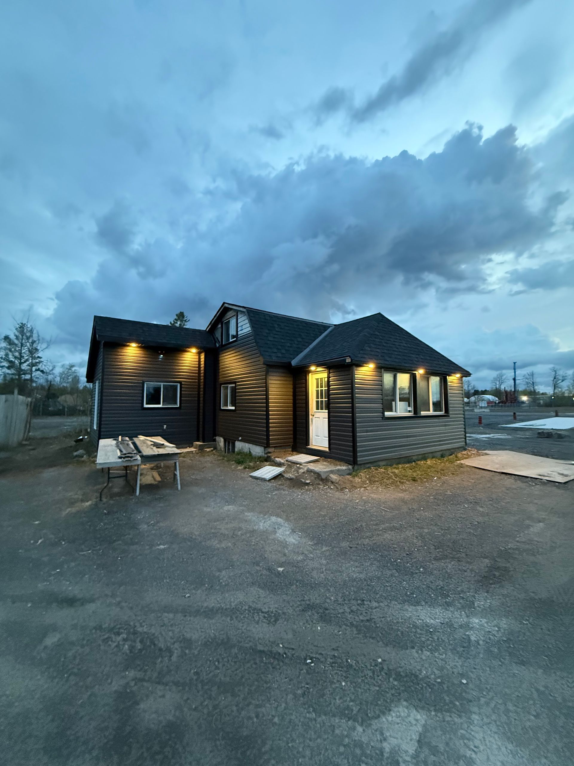 Dark-colored cabin with lit lights under a cloudy sky. Picnic table in front. Gravel ground.