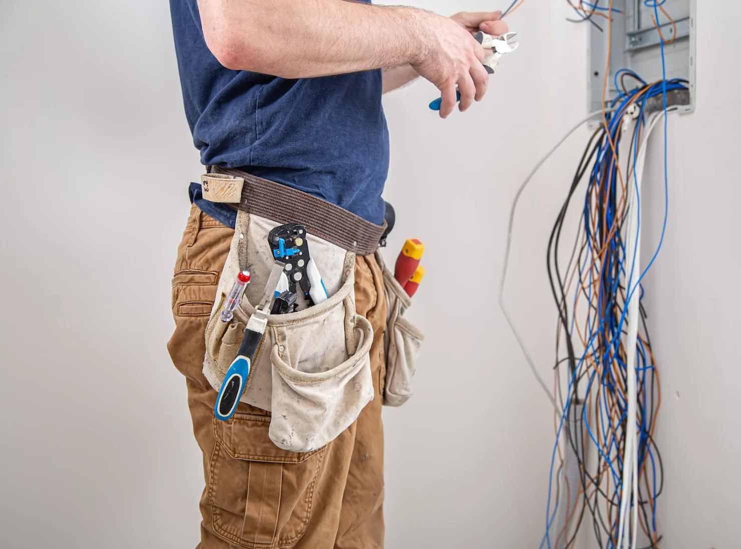 Electrician cutting wires near a wall-mounted panel.  Wires are blue, white, and brown. He wears a tool belt and brown pants.