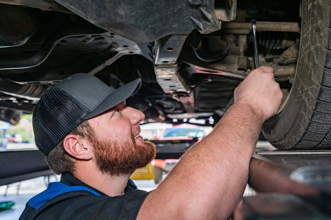 Mechanic inspecting a car’s undercarriage on a lift, wearing a cap and working with a tool