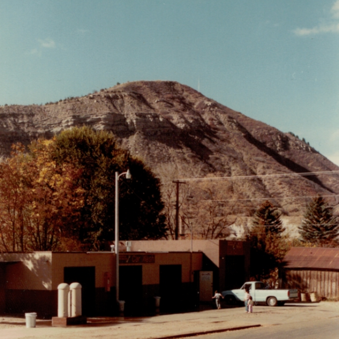 Desert roadside scene with a large rocky hill, trees, small buildings, and a parked car under a clear sky.