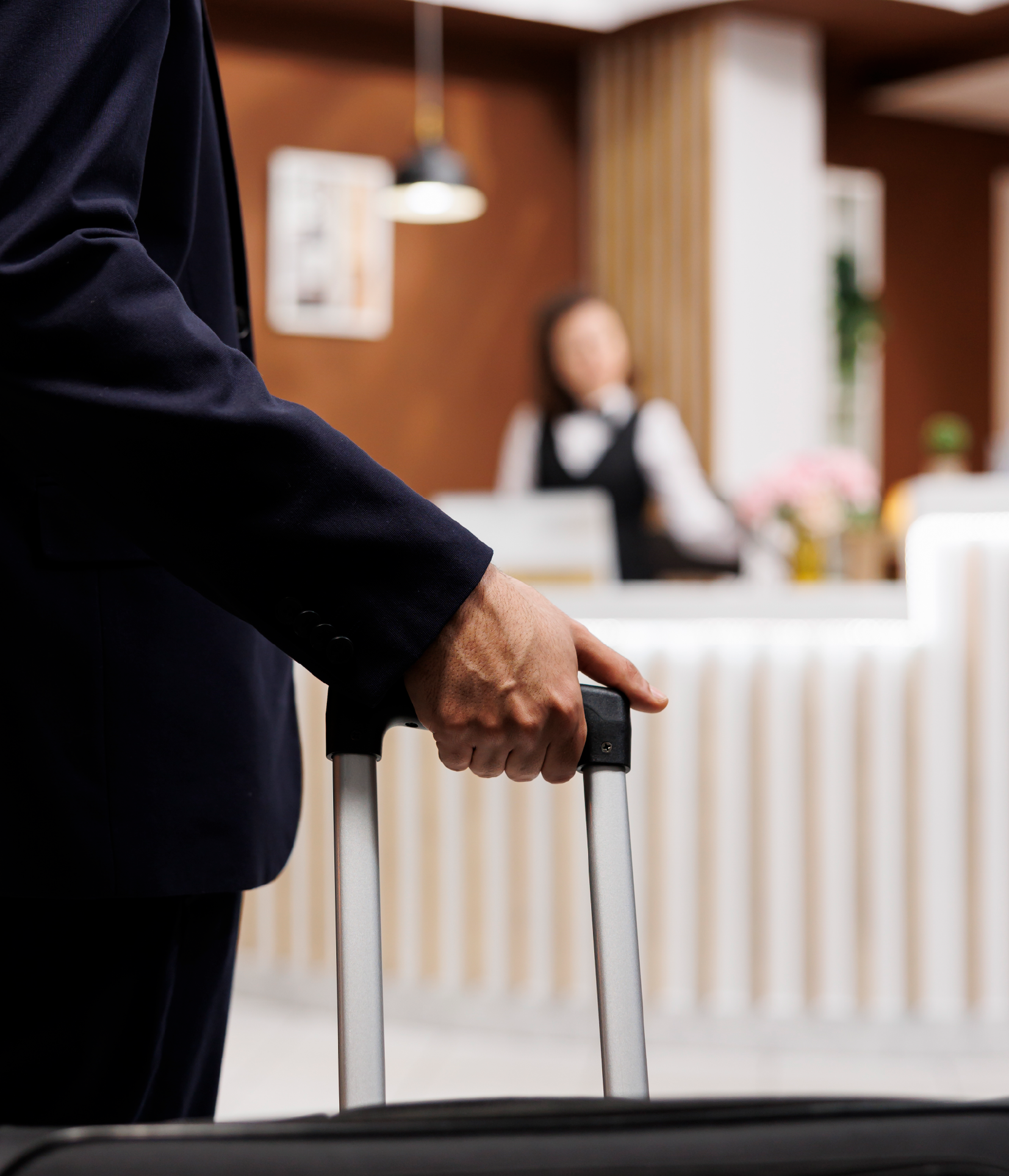 Una persona vestida con un traje oscuro sostiene el asa de una maleta en la recepción de un hotel, con un miembro del personal al fondo.