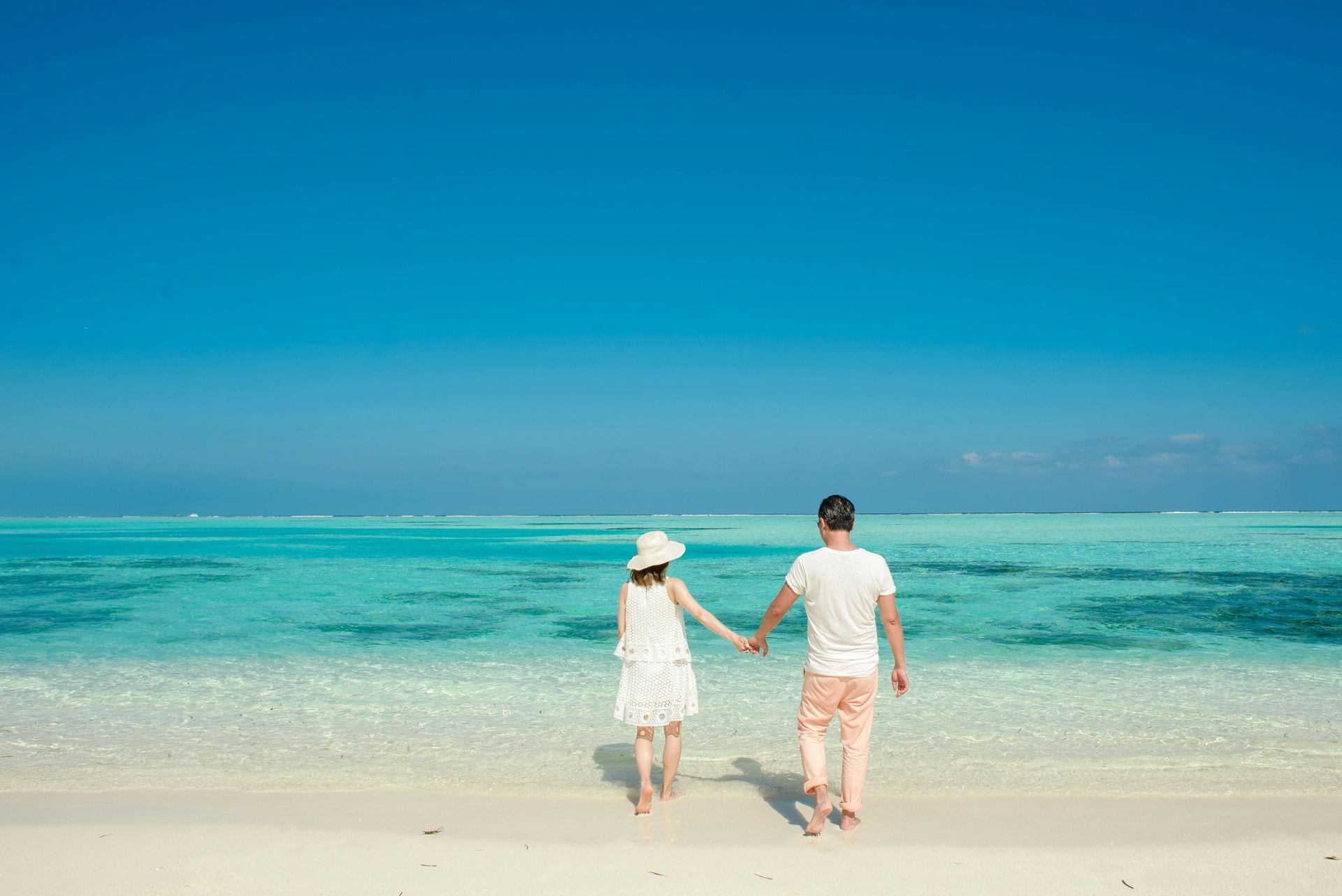 Una pareja camina de la mano hacia las aguas turquesas de una playa tropical bajo un cielo azul brillante.