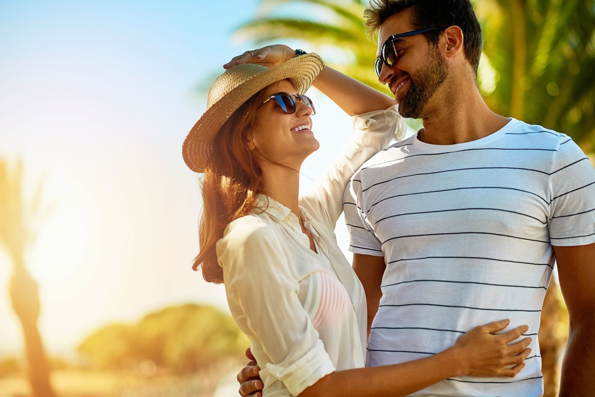 Una pareja con gafas de sol se abraza al aire libre bajo un sol tropical, mirándose y sonriendo.