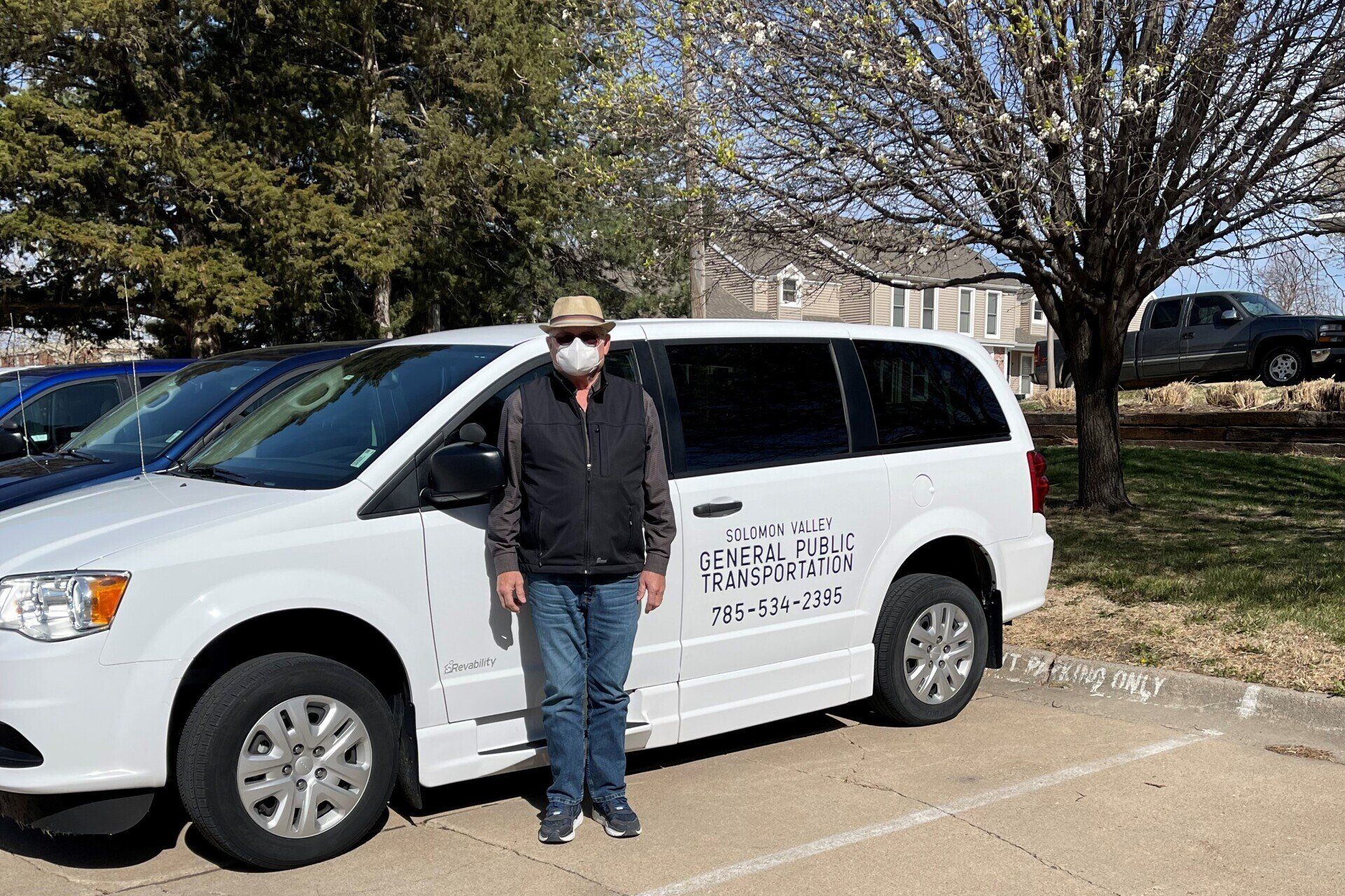 A white SVT mini van parked in a parking lot with trees in the background and a male driver standing in front