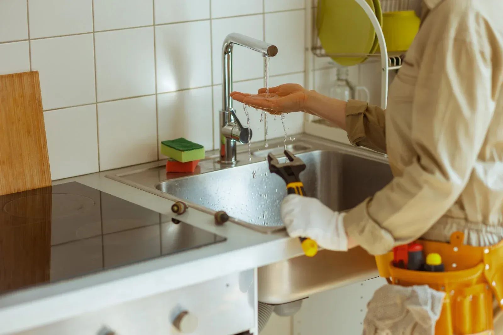 Person washing dishes at a kitchen sink with running water and cleaning supplies nearby
