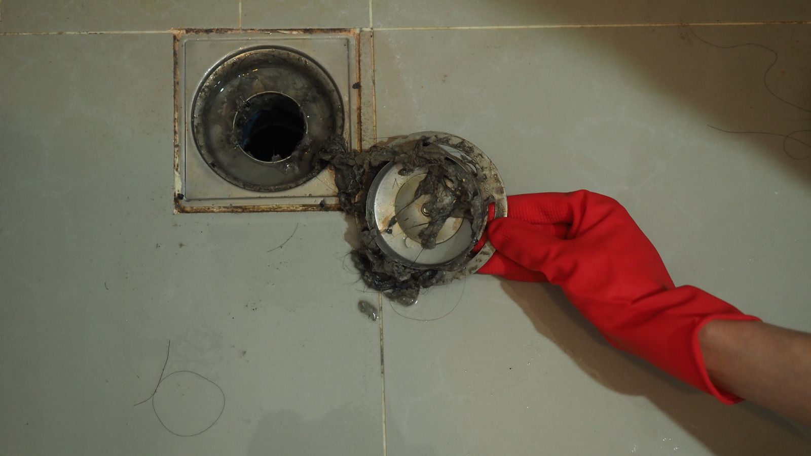 Gloved hand holding a metal drain cover over an open pipe hole in a tiled floor