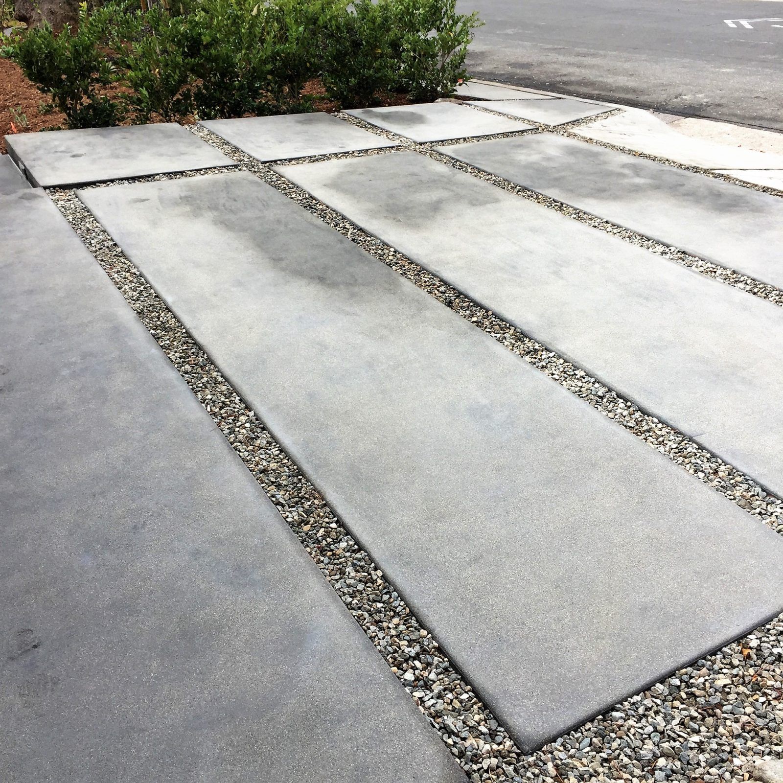 A concrete driveway with gravel and a bush in the background