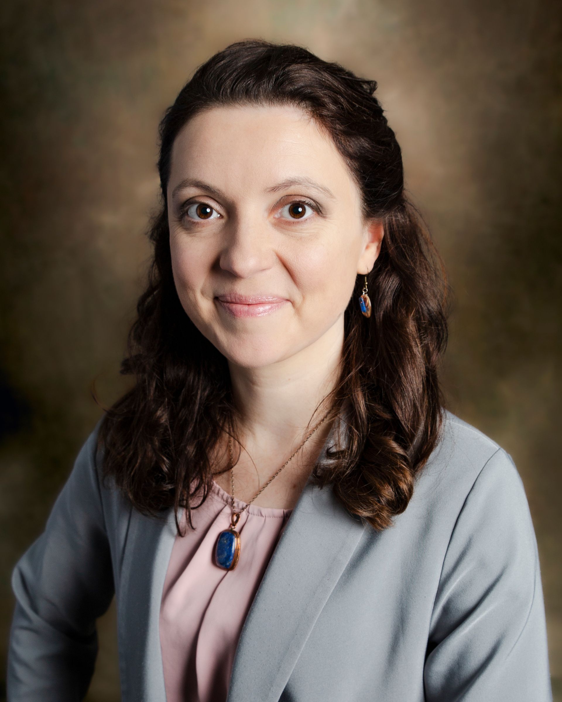 A woman with shoulder-length wavy brown hair wearing a blue blazer and a necklace, posed against a mottled brown backdrop.