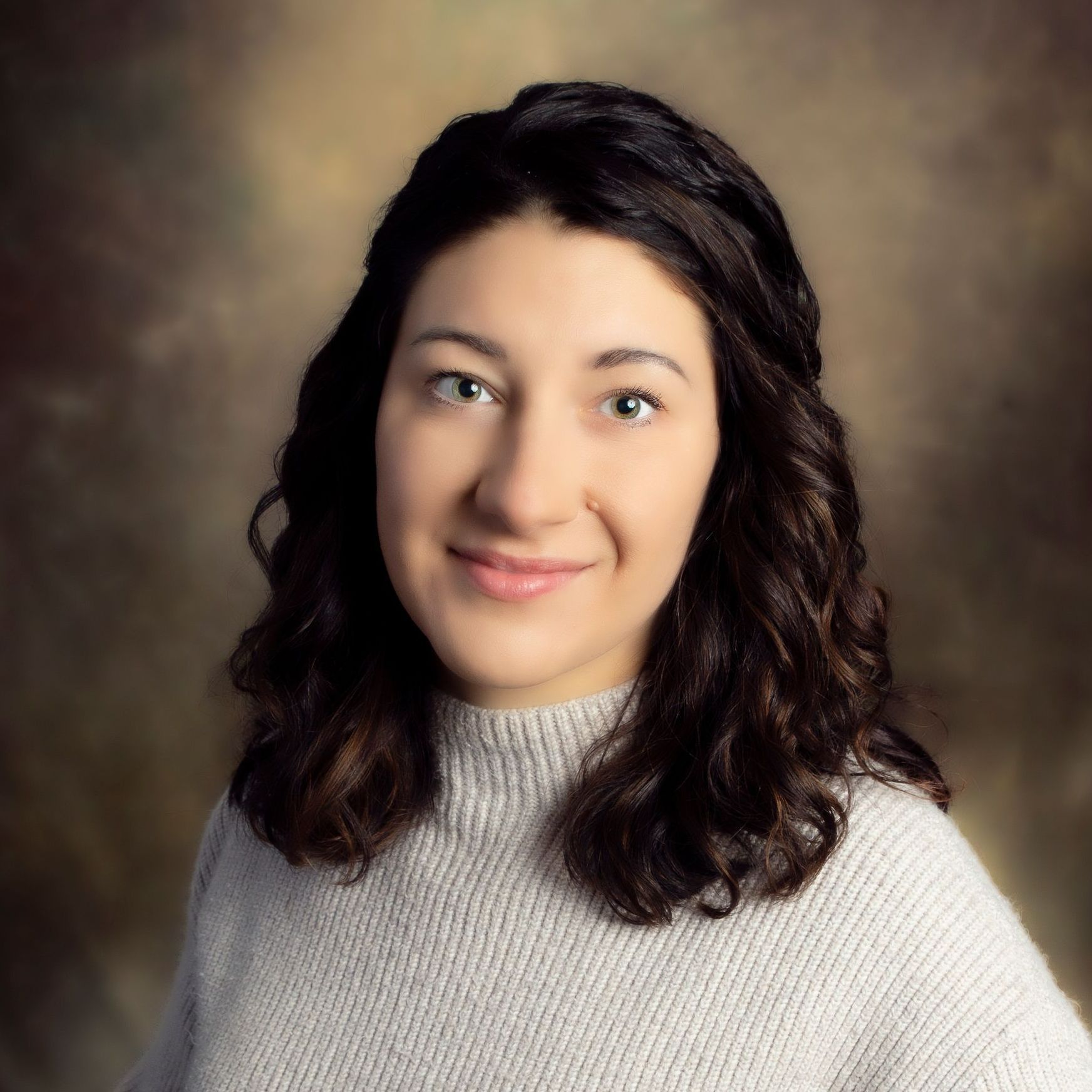 A person with long, wavy dark hair wears a light grey knit sweater against a warm, mottled brown background.