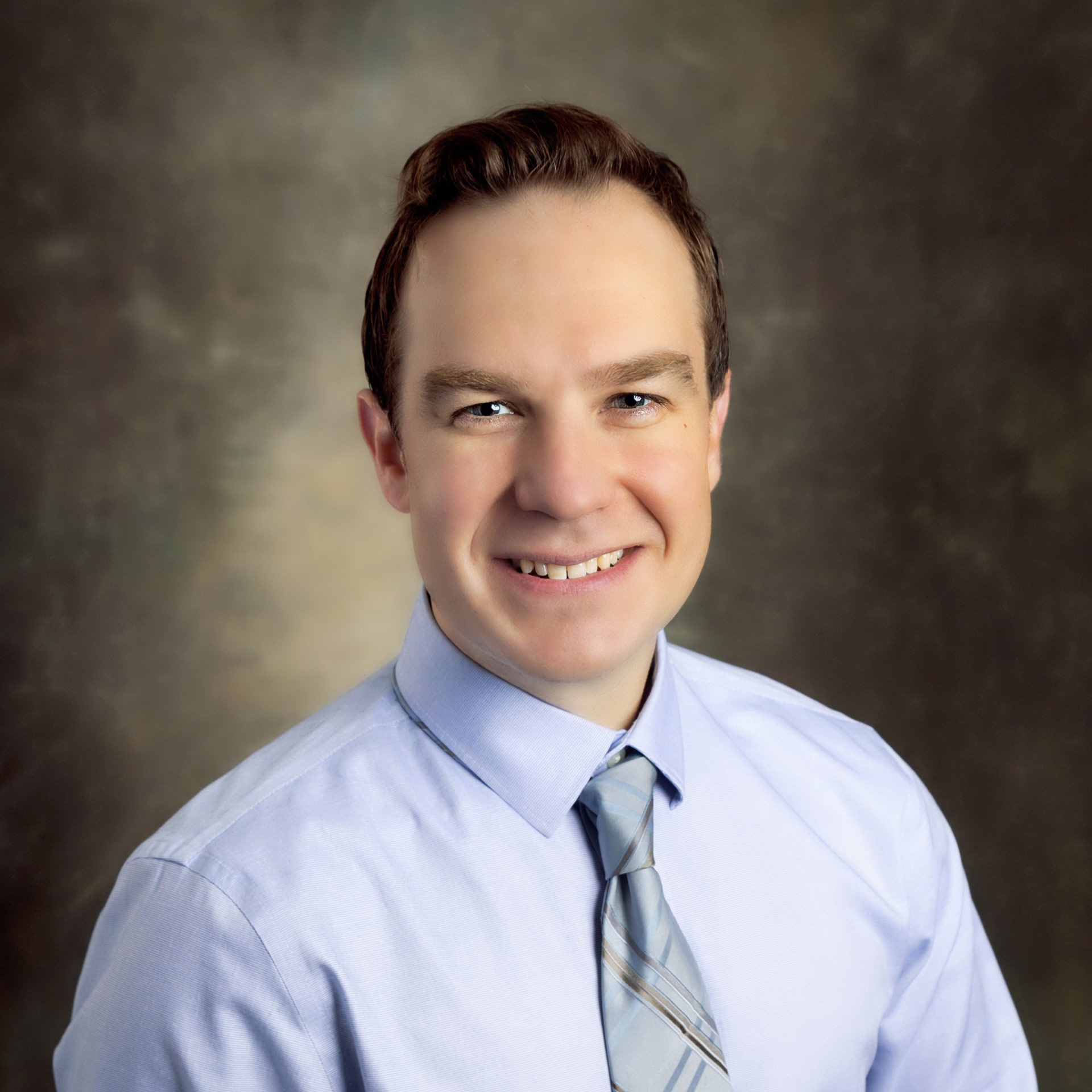 A person with short brown hair smiles while wearing a light blue button-down shirt and a patterned blue tie.