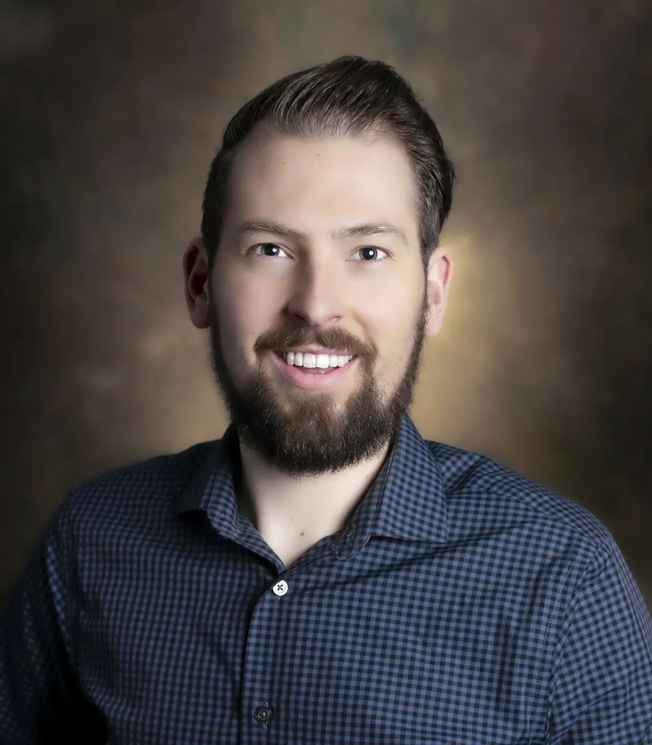 A smiling person with a short beard, wearing a blue patterned button-down shirt against a blurred brown background.