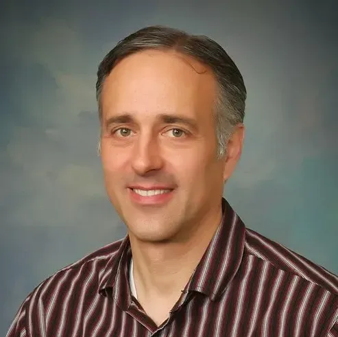 Headshot of a man with short brown hair smiling, wearing a dark striped button-down shirt against a blue background.