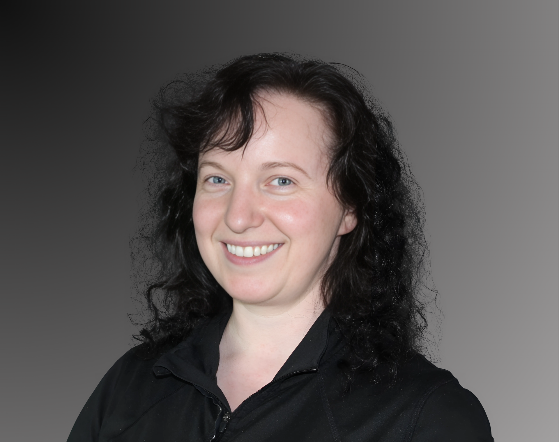 A person with dark, wavy hair and a friendly smile, wearing a black shirt against a grey gradient background.