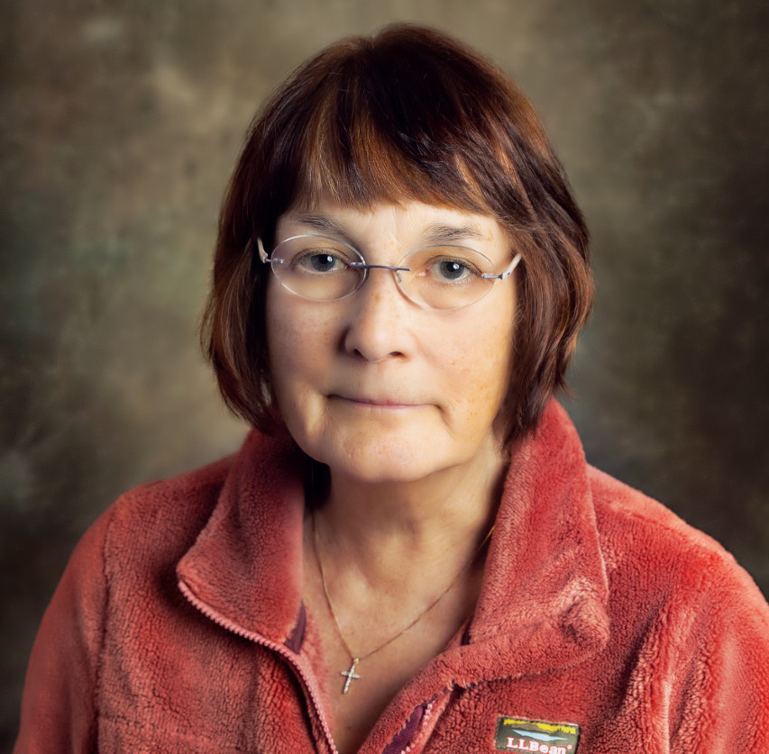A person with auburn hair and glasses wearing a red fleece jacket and a cross necklace against a dark, mottled background.