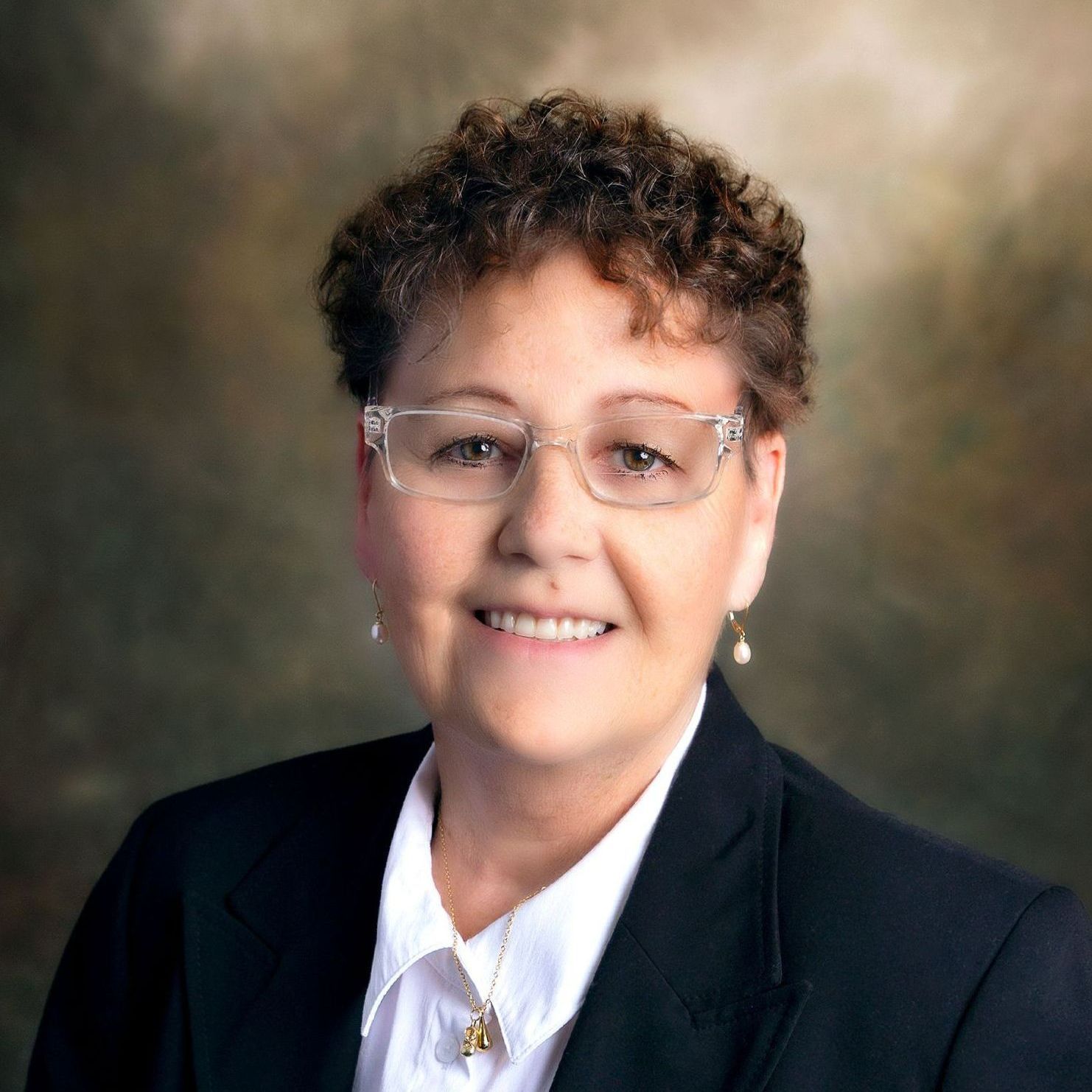 Professional portrait of a person with short curly hair wearing glasses, a white collared shirt, and a black blazer.