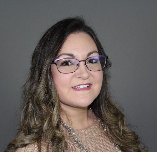 A smiling person with long brown hair and glasses wearing a textured beige sweater against a plain gray background.