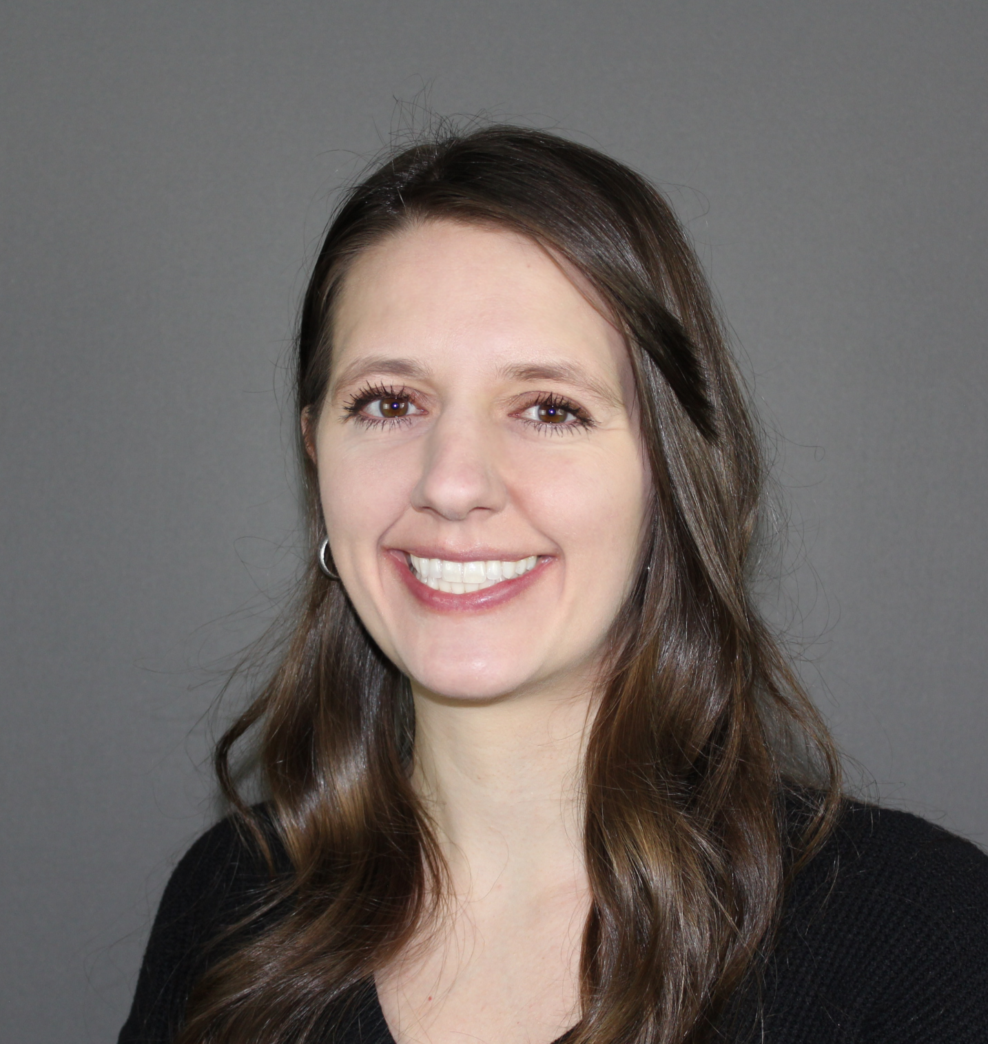A smiling person with long brown hair, wearing a black top against a solid grey background.