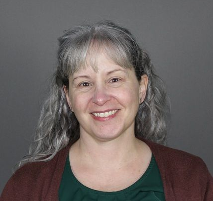 Smiling woman with long, wavy gray-streaked hair, wearing a dark green top under a maroon cardigan against a gray background.