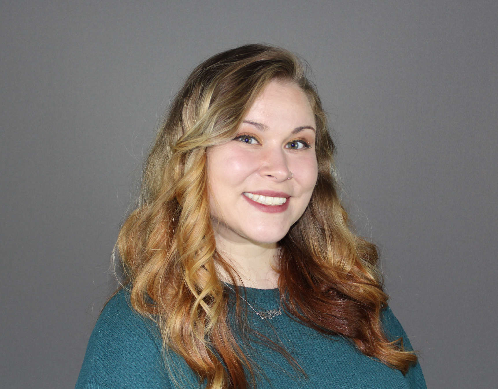 A person with long, wavy blonde and brown hair smiles warmly, wearing a teal sweater against a plain gray background.