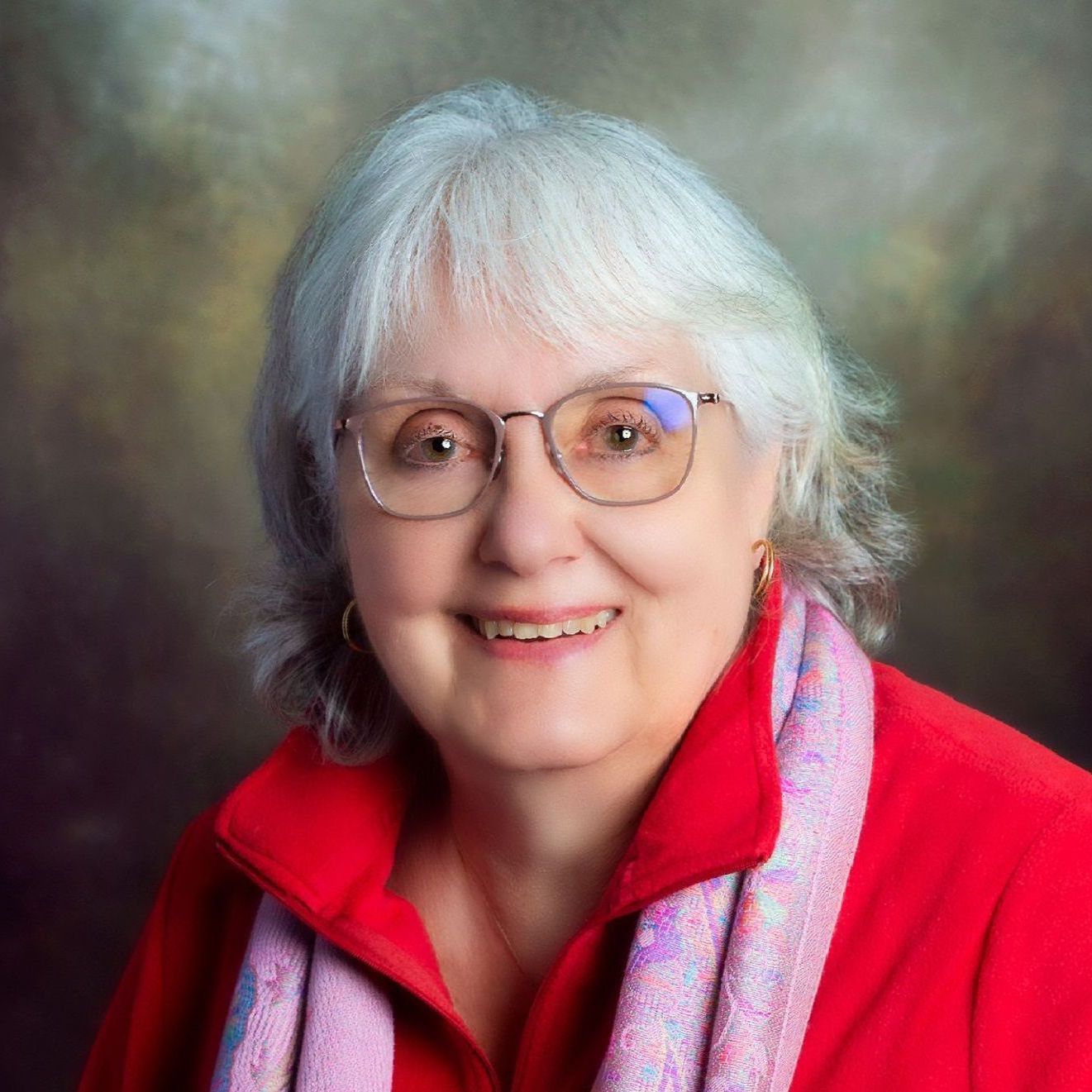 A smiling person with gray hair and glasses, wearing a bright red top and a patterned scarf against a dark background.