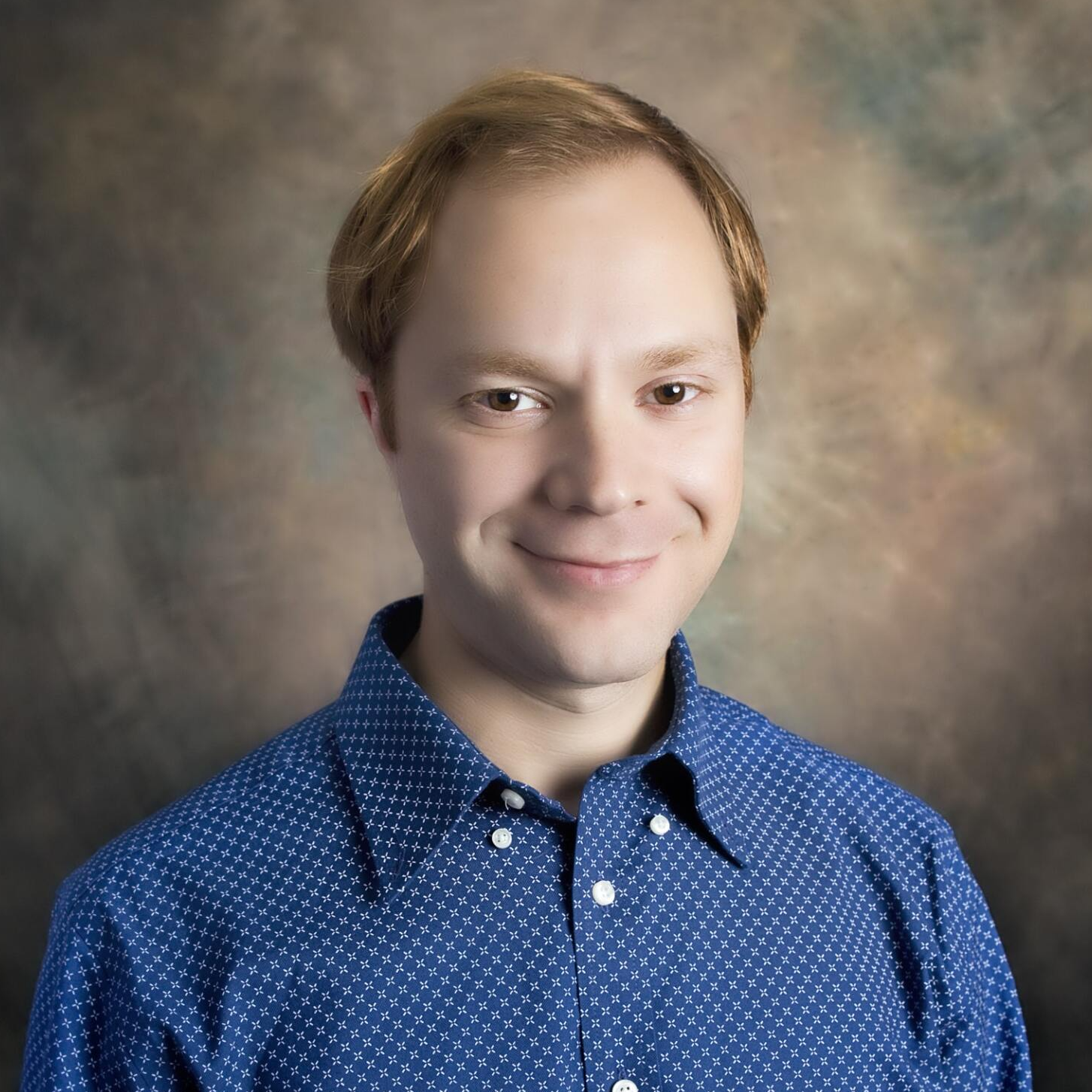Headshot of a smiling person with short light brown hair, wearing a blue button-down shirt against a mottled background.